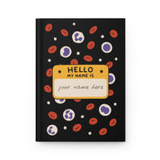 Black hardcover journal front cover with red and white blood cells and a yellow 'HELLO MY NAME IS' name tag reading 'your name here'