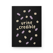 Front cover of a black hardcover journal with cream 'urine credible' lettering and lavender and cream crystals