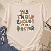 Garment-dyed unisex t-shirt in Bay printed with 'Yes, I'm Old Enough to Be a Doctor' — general medicine humor tee for young physicians.