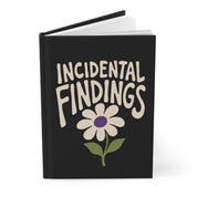 Black hardcover journal angled with pages visible, cream 'Incidental Findings' lettering above a daisy