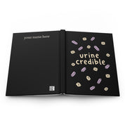 Black hardcover journal laid open flat, back cover 'your name here', front cover 'urine credible' with crystals