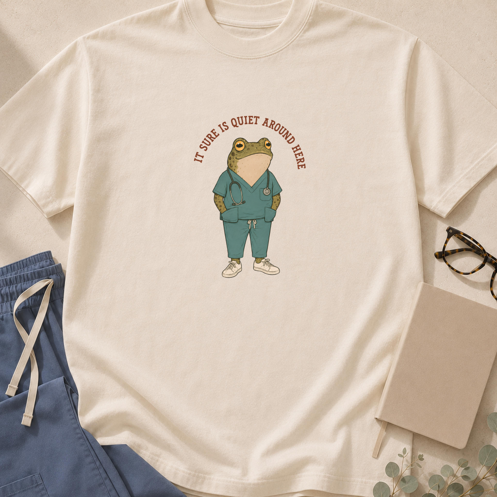 Garment-dyed unisex t-shirt in Bay printed with 'It Sure Is Quiet Around Here' beside a wide-eyed frog graphic — ER and ICU jinx humor tee for nurses and paramedics.