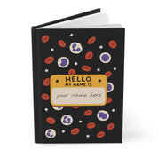 Black hardcover journal angled, cover open slightly, with blood cells and a 'HELLO MY NAME IS' tag reading 'your name here'