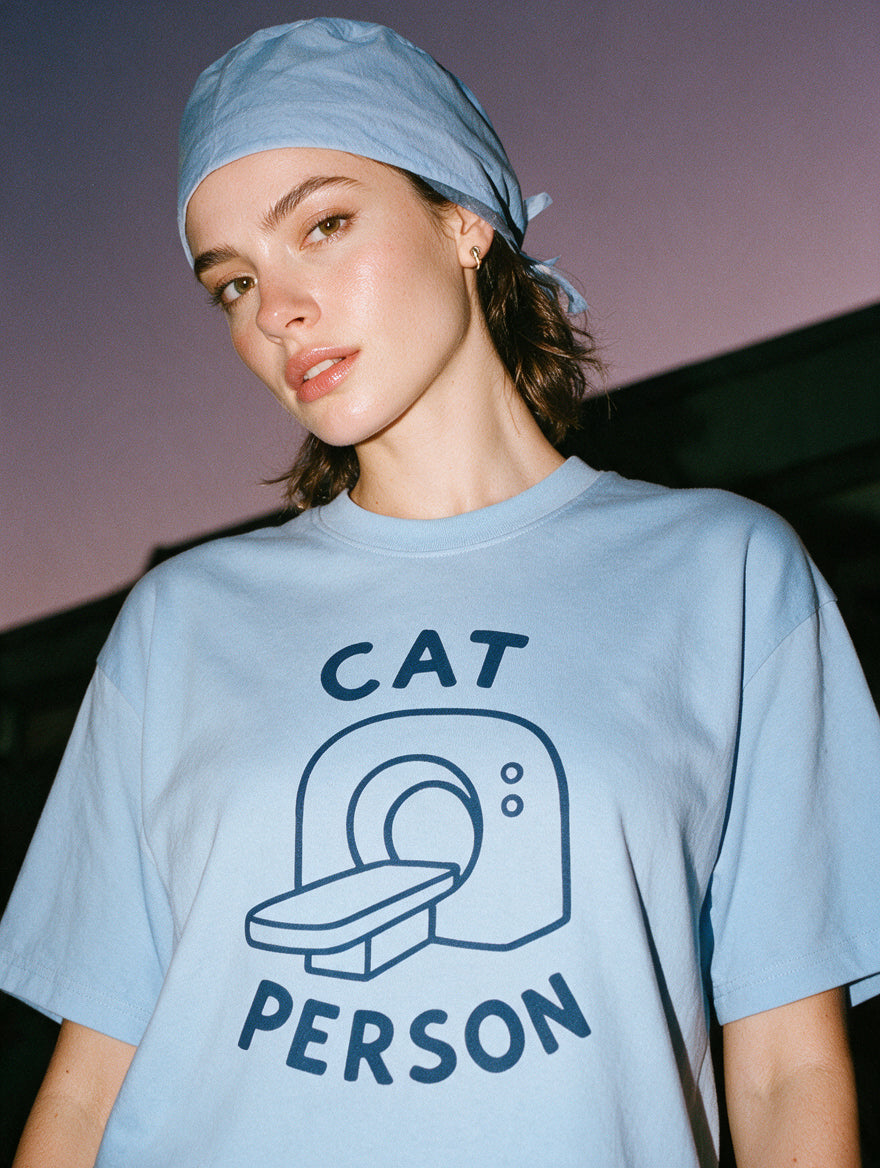 Woman in a scrub cap, photographed with flash at night, wearing a light blue cotton T-shirt with a hand-drawn CT scanner and the words "Cat Person."
