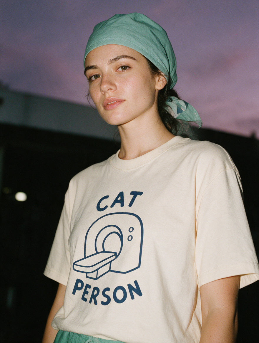 Woman in a scrub cap, photographed with flash at night, wearing a cream cotton T-shirt with a hand-drawn CT scanner and the words "Cat Person."