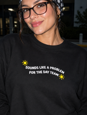 Model wearing the black embroidered 'Sounds Like a Problem for the Day Team' sweatshirt, daytime editorial flash photo outside a hospital.