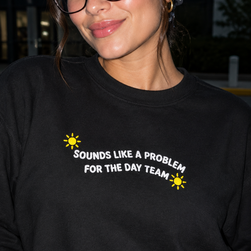Model wearing the black embroidered 'Sounds Like a Problem for the Day Team' sweatshirt, daytime editorial flash photo outside a hospital.