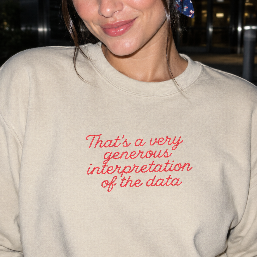 Model wearing the sand embroidered 'Generous Interpretation of the Data' sweatshirt, daytime editorial flash photo outside a hospital.