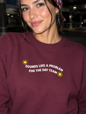 Model wearing the maroon embroidered 'Sounds Like a Problem for the Day Team' sweatshirt, daytime editorial flash photo outside a hospital.