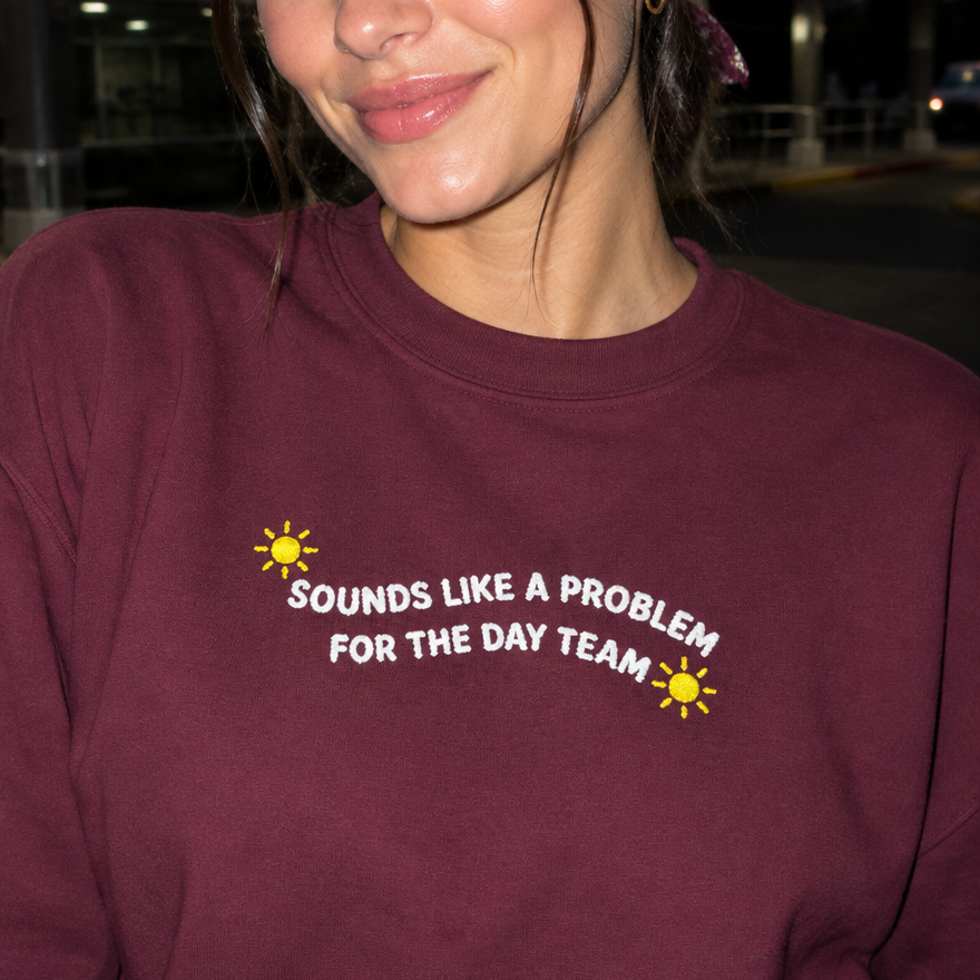 Model wearing the maroon embroidered 'Sounds Like a Problem for the Day Team' sweatshirt, daytime editorial flash photo outside a hospital.