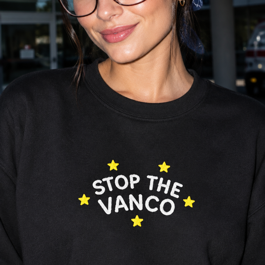 Model wearing the black embroidered 'Stop the Vanco' sweatshirt, daytime editorial flash photo outside a hospital.