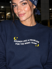 Model wearing the navy embroidered 'Sounds Like a Problem for the Night Team' sweatshirt, daytime editorial flash photo outside a hospital.
