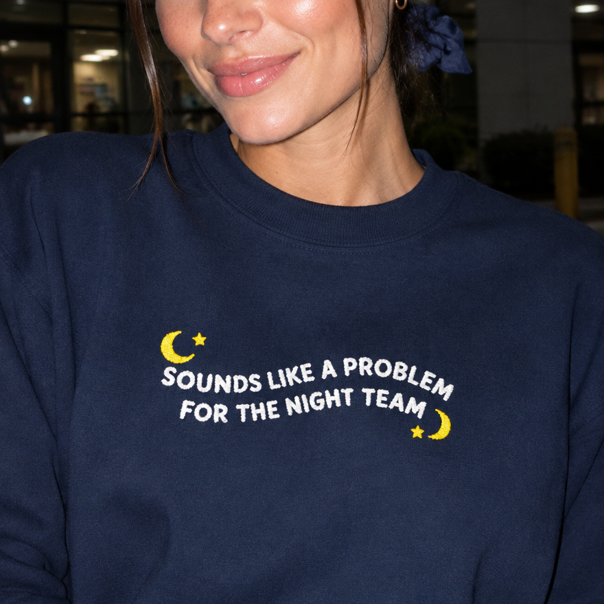 Model wearing the navy embroidered 'Sounds Like a Problem for the Night Team' sweatshirt, daytime editorial flash photo outside a hospital.