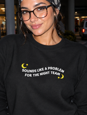 Model wearing the black embroidered 'Sounds Like a Problem for the Night Team' sweatshirt, daytime editorial flash photo outside a hospital.