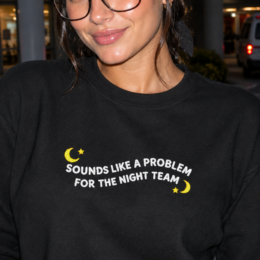 Model wearing the black embroidered 'Sounds Like a Problem for the Night Team' sweatshirt, daytime editorial flash photo outside a hospital.