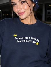 Model wearing the navy embroidered 'Sounds Like a Problem for the Day Team' sweatshirt, daytime editorial flash photo outside a hospital.