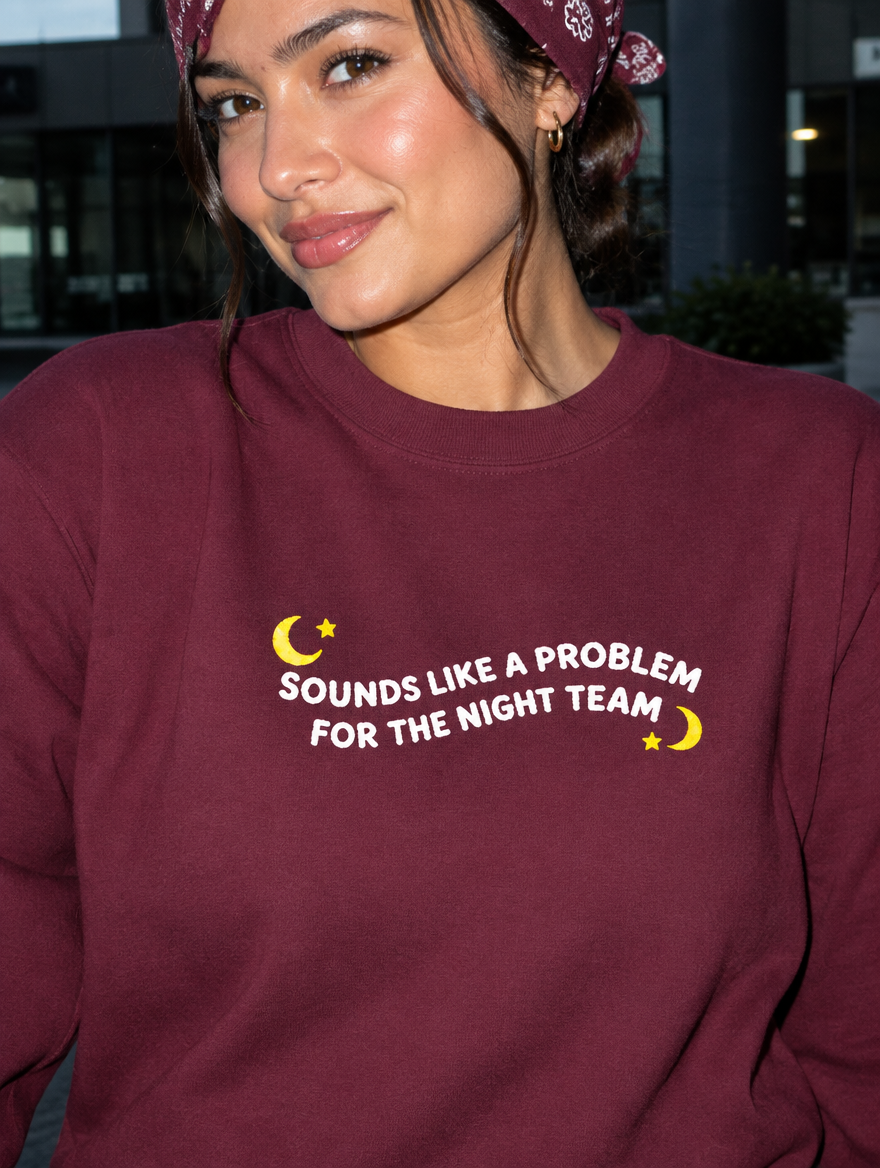 Model wearing the maroon embroidered 'Sounds Like a Problem for the Night Team' sweatshirt, daytime editorial flash photo outside a hospital.