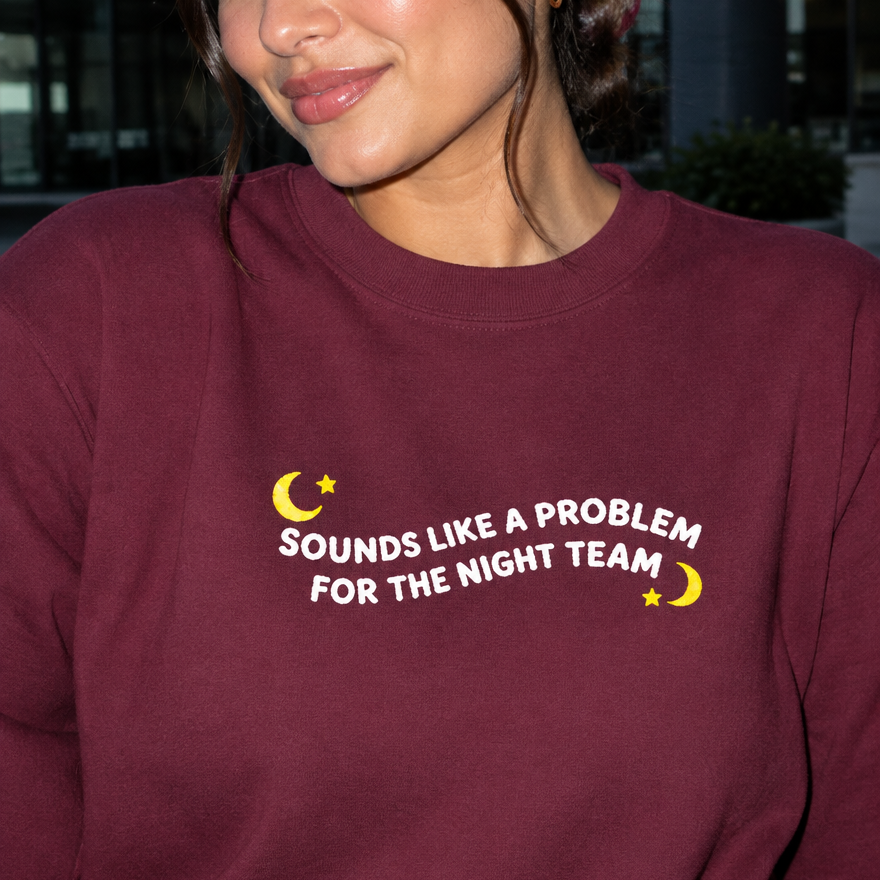 Model wearing the maroon embroidered 'Sounds Like a Problem for the Night Team' sweatshirt, daytime editorial flash photo outside a hospital.