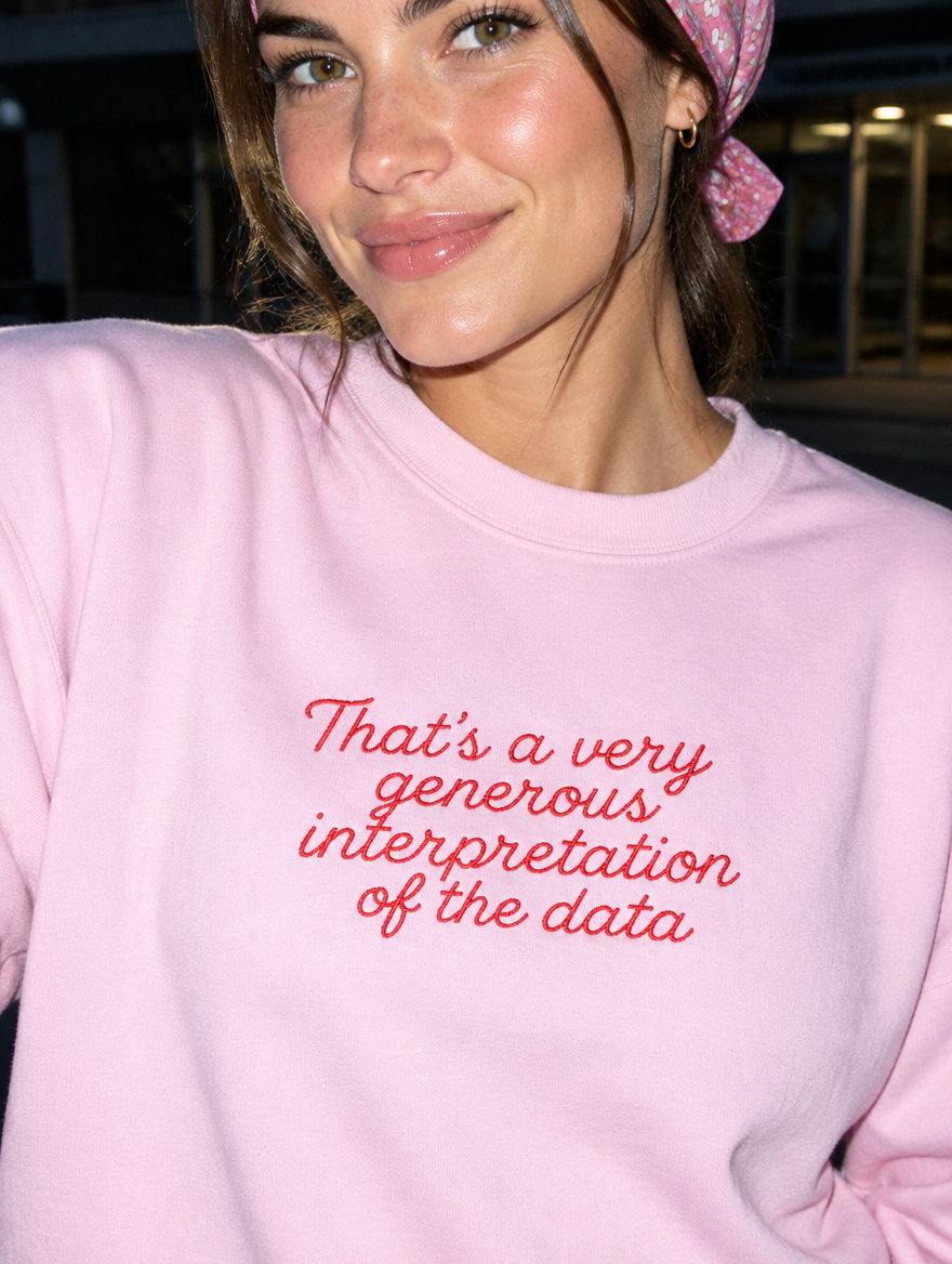 Model wearing the light pink embroidered 'Generous Interpretation of the Data' sweatshirt, daytime editorial flash photo outside a hospital.