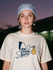 Woman photographed with flash at night, wearing an apricot cotton T-shirt with stork carrying a bundle with the words 'On Stork Duty'.