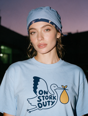 Woman photographed with flash at night, wearing a light blue cotton T-shirt with stork carrying a bundle with the words 'On Stork Duty'.