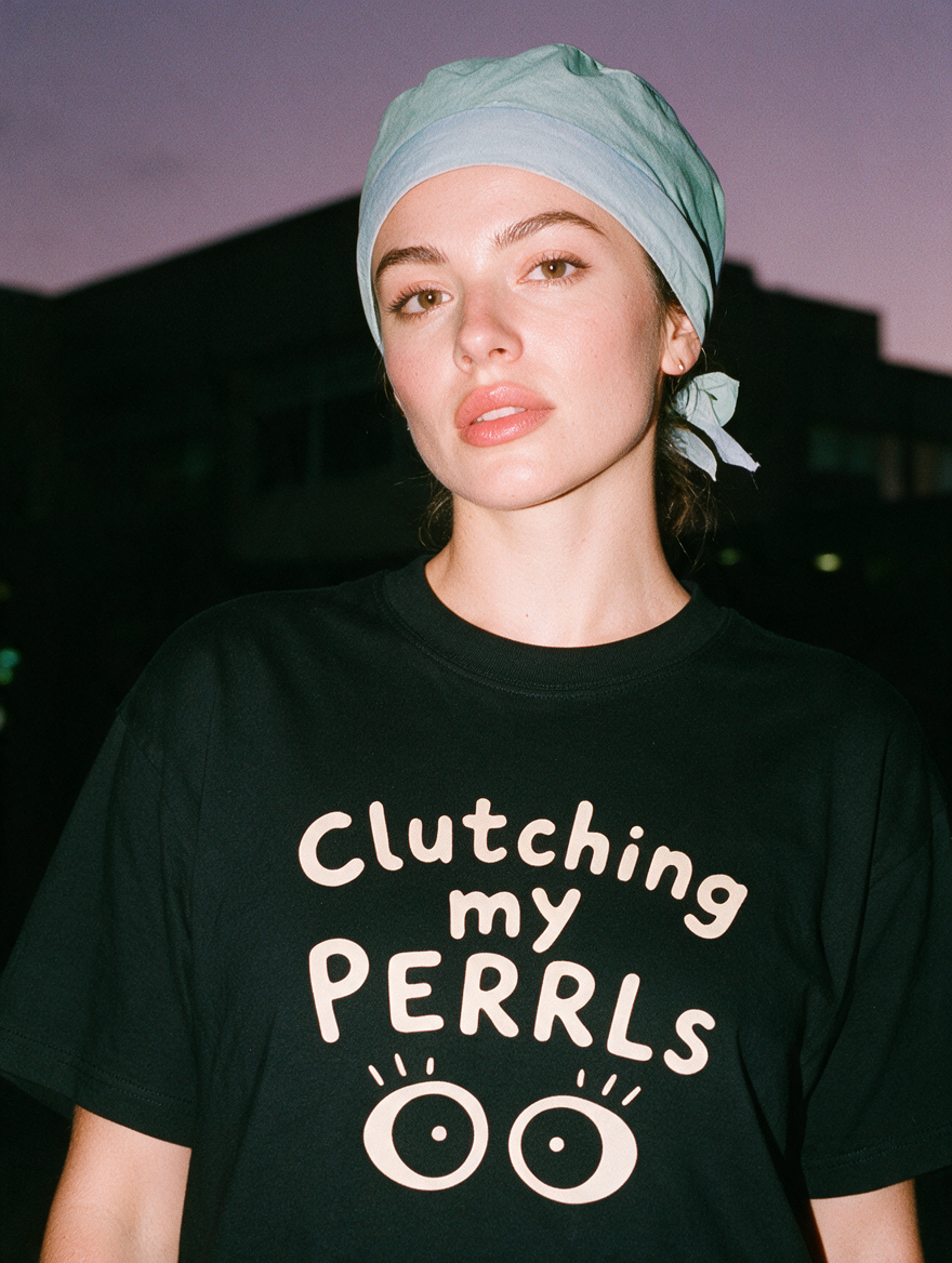 Woman photographed with flash at night, wearing a black cotton T-shirt with two wide round eyes under the words 'Clutching my PERRLs'.