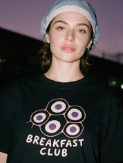 Woman in a scrub cap, photographed with flash at night, wearing a black cotton T-shirt with a "Breakfast Club" design.