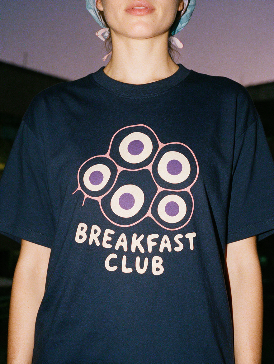 Woman in a scrub cap, photographed with flash at night, wearing a navy blue cotton T-shirt with a "Breakfast Club" design.
