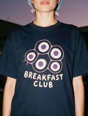 Woman in a scrub cap, photographed with flash at night, wearing a navy blue cotton T-shirt with a "Breakfast Club" design.