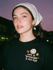 Woman photographed with flash at night, wearing a black cotton T-shirt with a smiley face, heart, and star with the words 'not a "provider," k thx bye'.