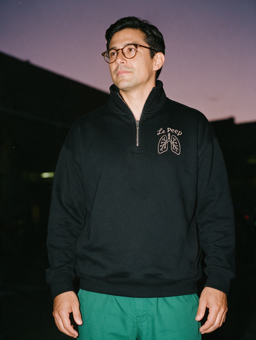 Man photographed with flash at night, wearing a black quarter-zip sweatshirt with the Le Peep design.