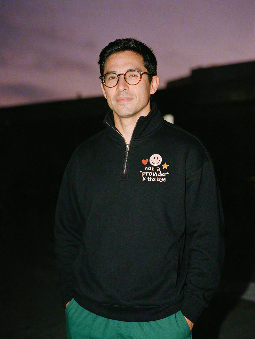 Man in round tortoiseshell glasses and green scrub pants, flash at night, wearing a black quarter-zip with a smiley, heart and star above 'not a "provider" k thx bye'