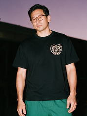 Man in round tortoiseshell glasses and green scrub pants, flash at night, wearing a black cotton T-shirt with a heart graphic 'Uptitrate GDMT for HFrEF as tolerated'