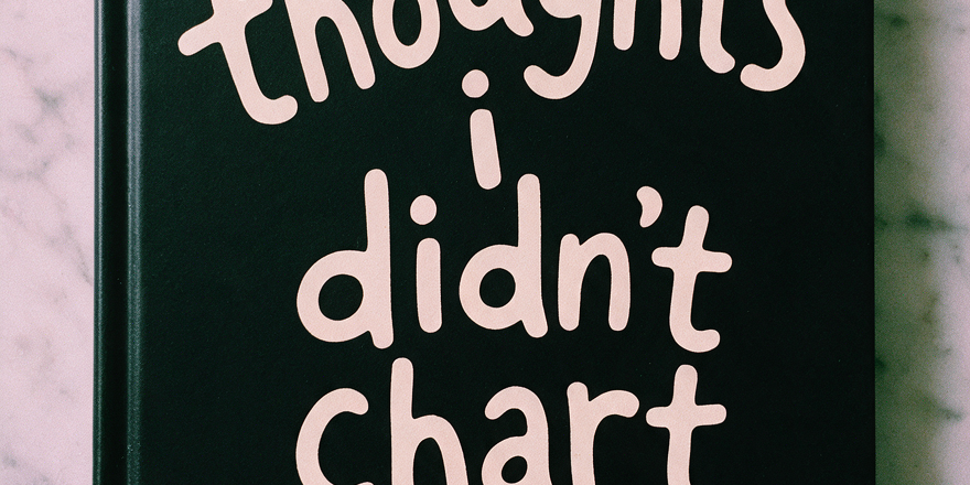 Black hardcover journal with cream lettering reading 'thoughts i didn't chart' on a marble surface
