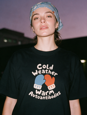 Woman photographed with flash at night, wearing a black cotton T-shirt with mittens and the text 'Cold Weather Warm Autoantibodies.'
