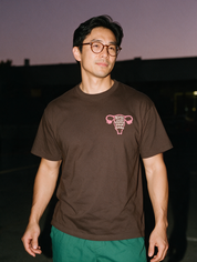 Man in tortoiseshell glasses, photographed with flash at night, wearing a coffee brown cotton T-shirt with a pink uterus and 'girls just wanna have fundus'