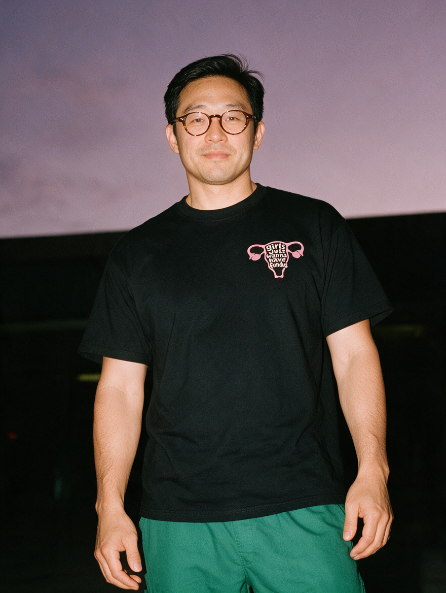 Man in tortoiseshell glasses, photographed with flash at night, wearing a black cotton T-shirt with a pink uterus and 'girls just wanna have fundus'