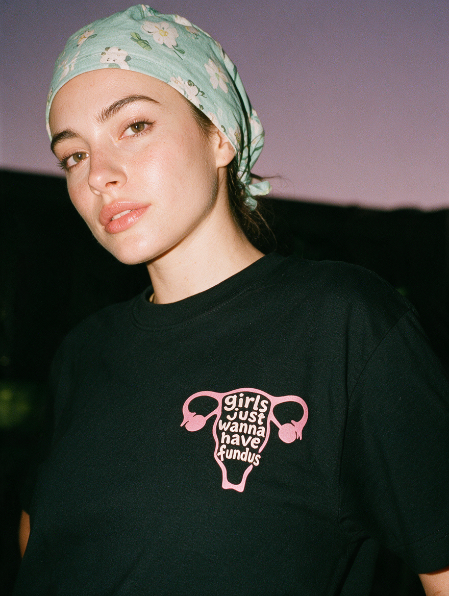 Woman in a scrub cap, photographed with flash at night, wearing a black cotton T-shirt with a pink uterus and 'girls just wanna have fundus'