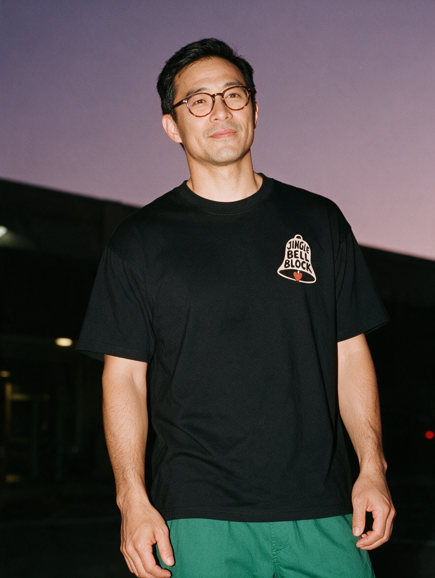 Man in glasses, photographed with flash at night, wearing a black cotton T-shirt with a small chest bell lettered "Jingle Bell Block"