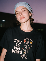 Woman photographed with flash at night wearing a black cotton T-shirt with 'Joy to the Ward' beside an IV pole wrapped in holiday lights