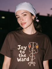 Woman photographed with flash at night wearing a coffee-brown cotton T-shirt with 'Joy to the Ward' beside an IV pole wrapped in holiday lights