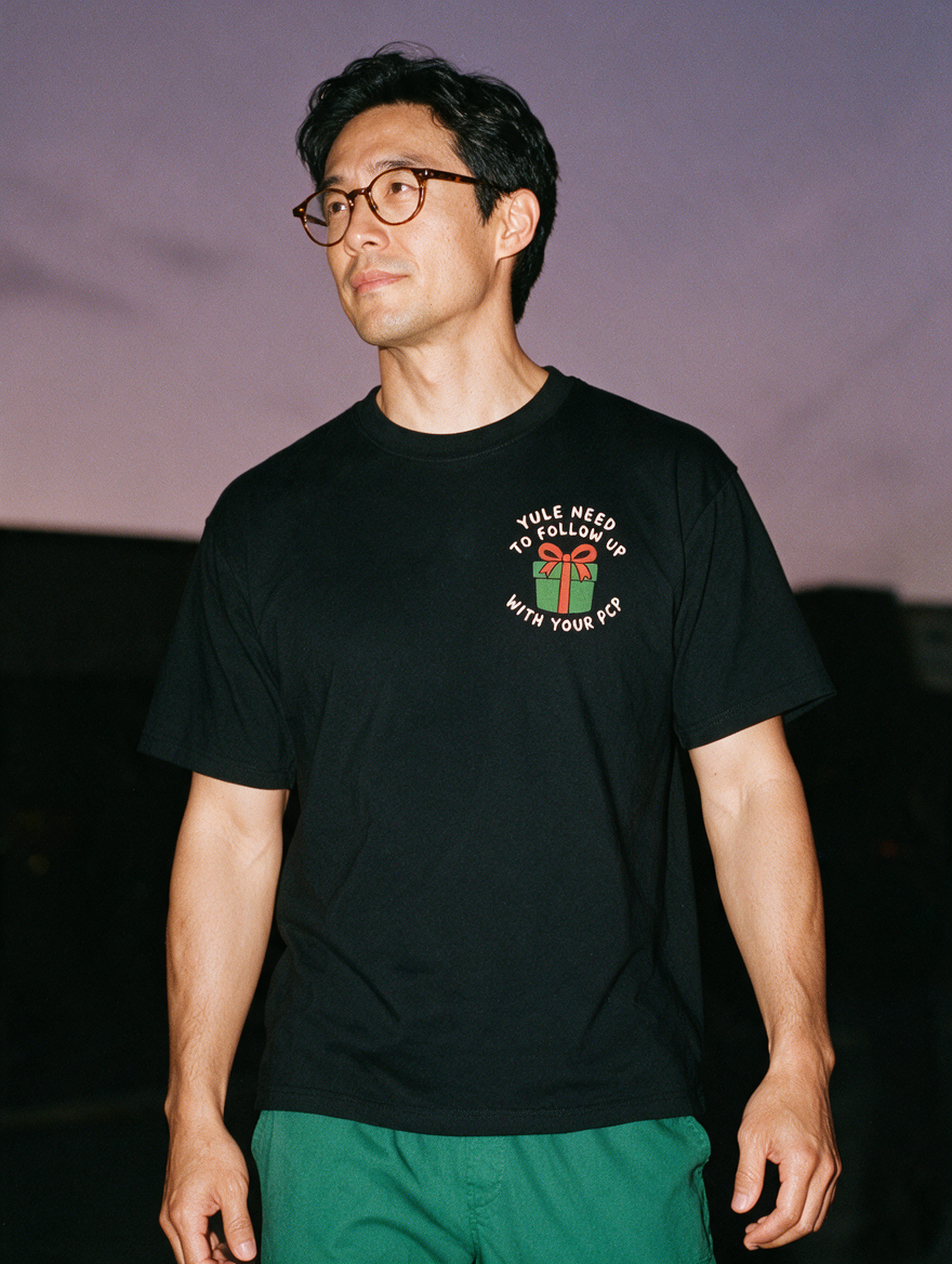Man in tortoiseshell glasses photographed with flash at night, wearing a black cotton T-shirt with a small green gift box and 'YULE NEED TO FOLLOW UP WITH YOUR PCP'