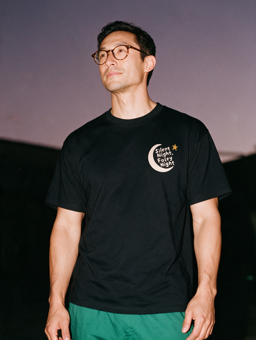 Man in tortoiseshell glasses photographed with flash at night, wearing a black cotton T-shirt with a crescent moon, a star and 'Silent Night, Foley Night'