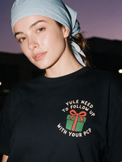 Woman photographed with flash at night, wearing a black cotton T-shirt with a small green gift box and 'YULE NEED TO FOLLOW UP WITH YOUR PCP'