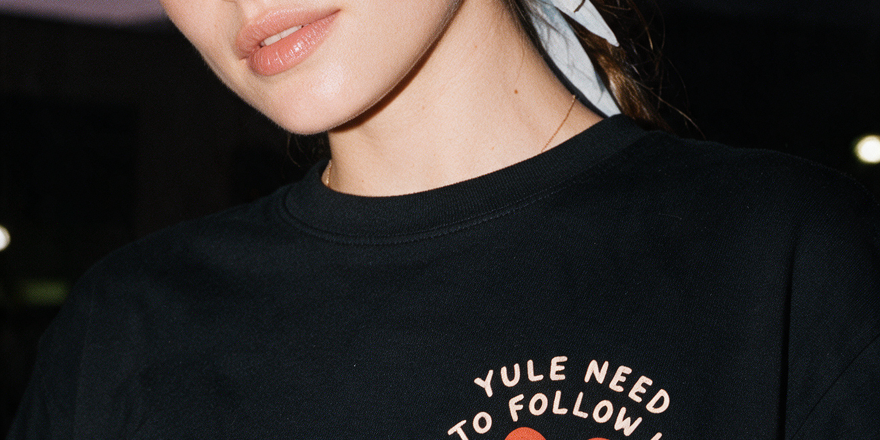 Woman photographed with flash at night, wearing a black cotton T-shirt with a small green gift box and 'YULE NEED TO FOLLOW UP WITH YOUR PCP'