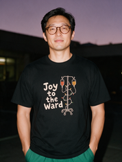 Man in glasses photographed with flash at night wearing a black cotton T-shirt with 'Joy to the Ward' beside an IV pole wrapped in holiday lights
