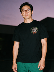 Man in tortoiseshell glasses, photographed with flash at night, wearing a black cotton T-shirt with a small wreath and the text 'Have Yourself a Merry Little Discharge.'