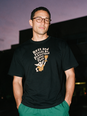 Man in round glasses and green scrub pants, photographed with flash at night, wearing a black cotton T-shirt printed 'Rest and Fluids Be Prescribed Unto Thee.'