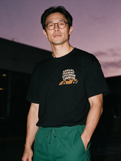 Man in round glasses and green scrub pants, photographed with flash at night, wearing a black cotton T-shirt with 'Annual Training Speedrun Champion' and an orange race car.
